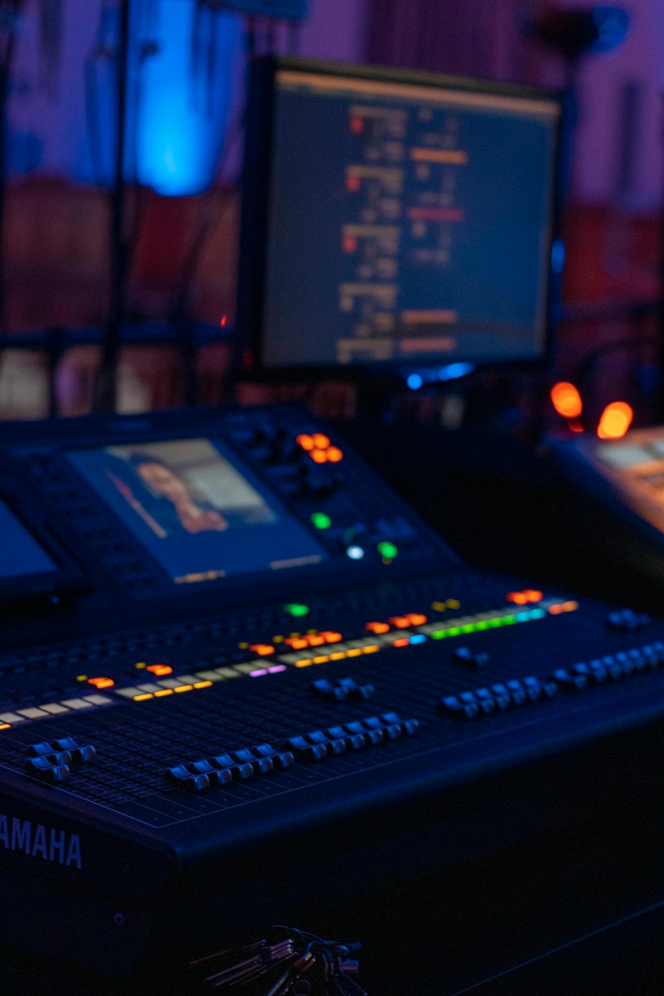 Close-up of a professional audio mixing console in a dimly lit music studio with monitors displaying audio software.